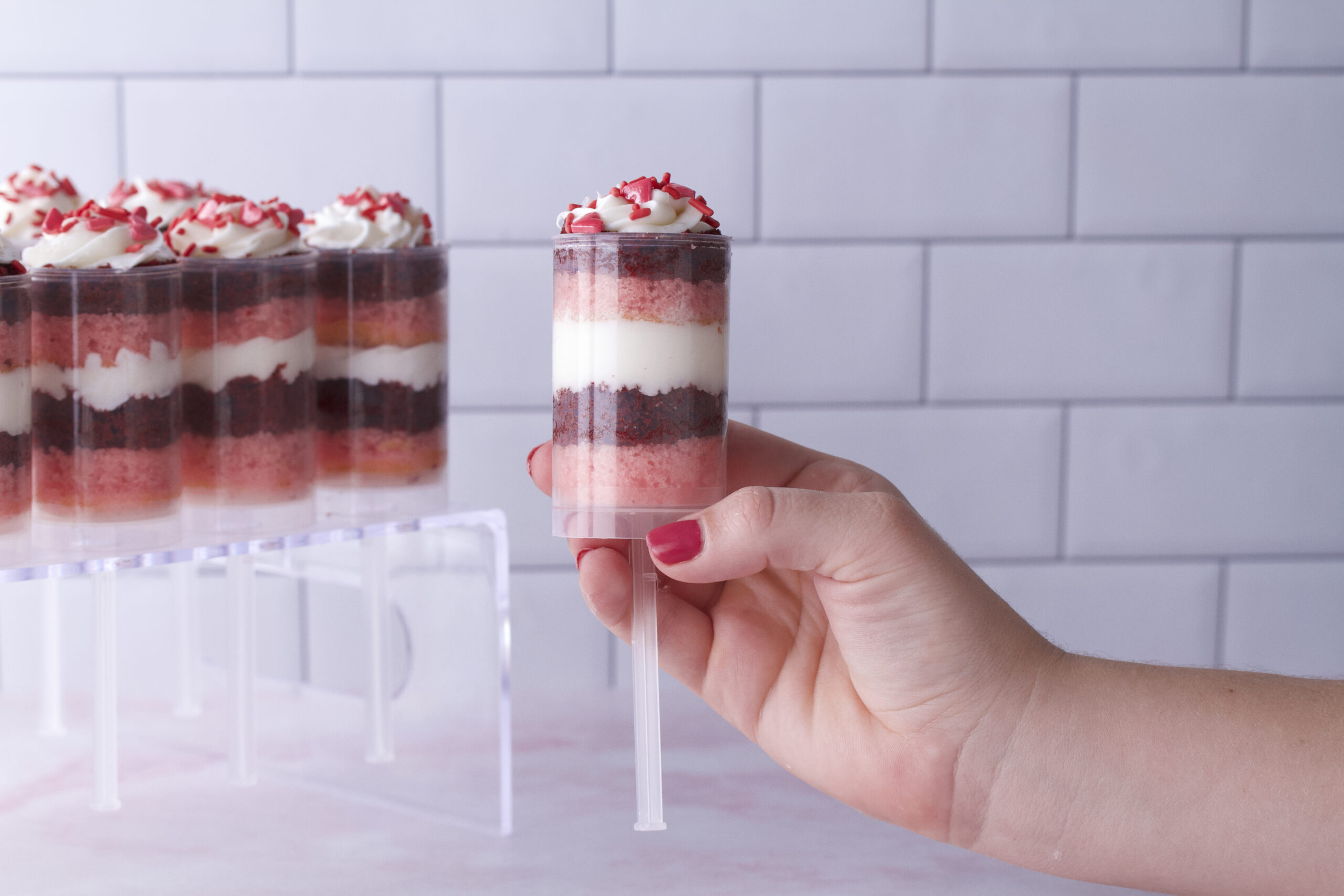 A hand holding a Berry Velvet Bliss cake push-up, showcasing layers of red velvet cake, strawberry cake, and buttercream frosting, topped with colorful Valentine’s Day sprinkles. Additional cake push-ups are arranged in the background, creating a festive dessert display.